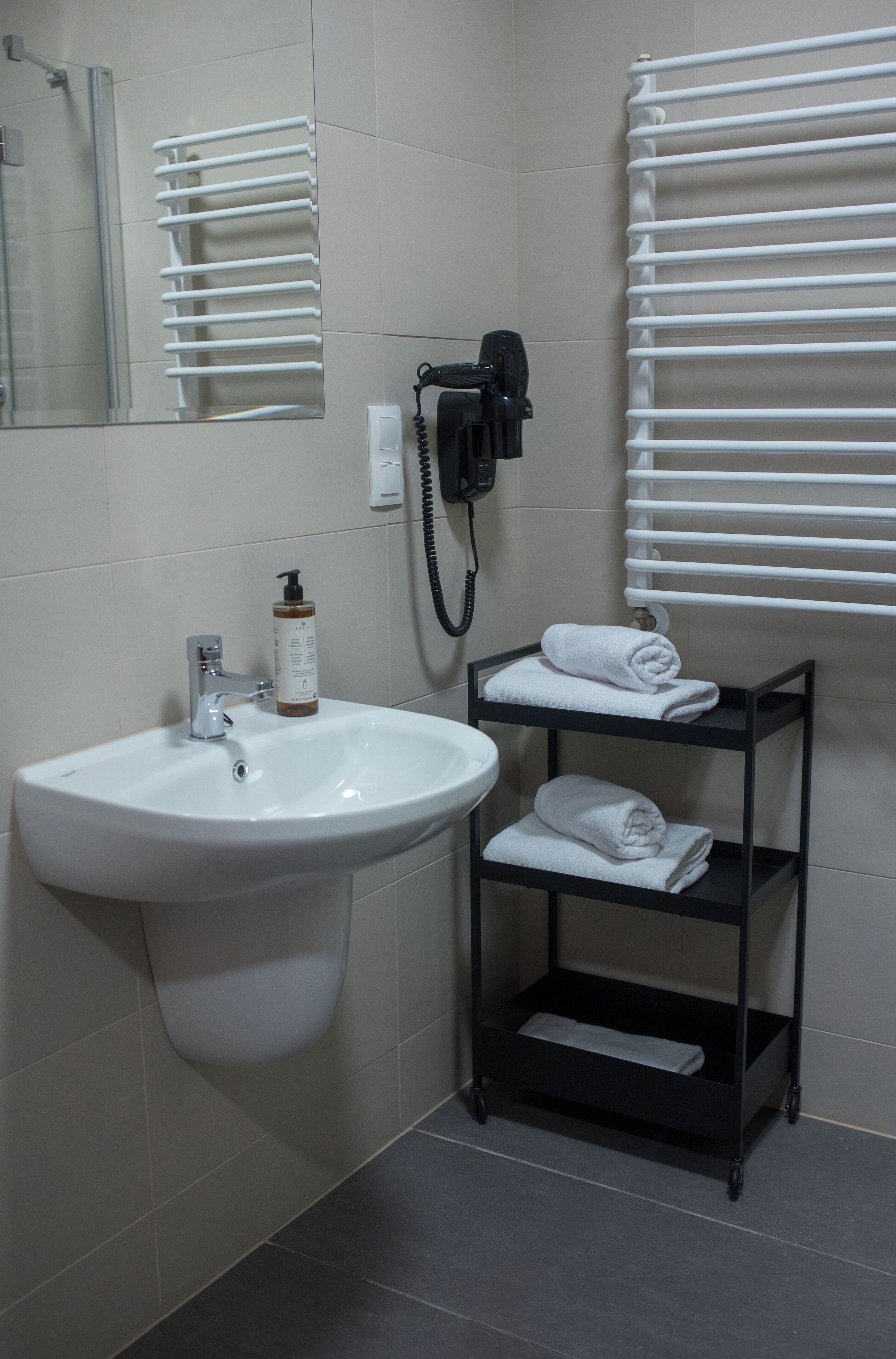 A bathroom with a white sink, a mirror, a towel rack, a shelf with towels, and a bottle of shampoo.