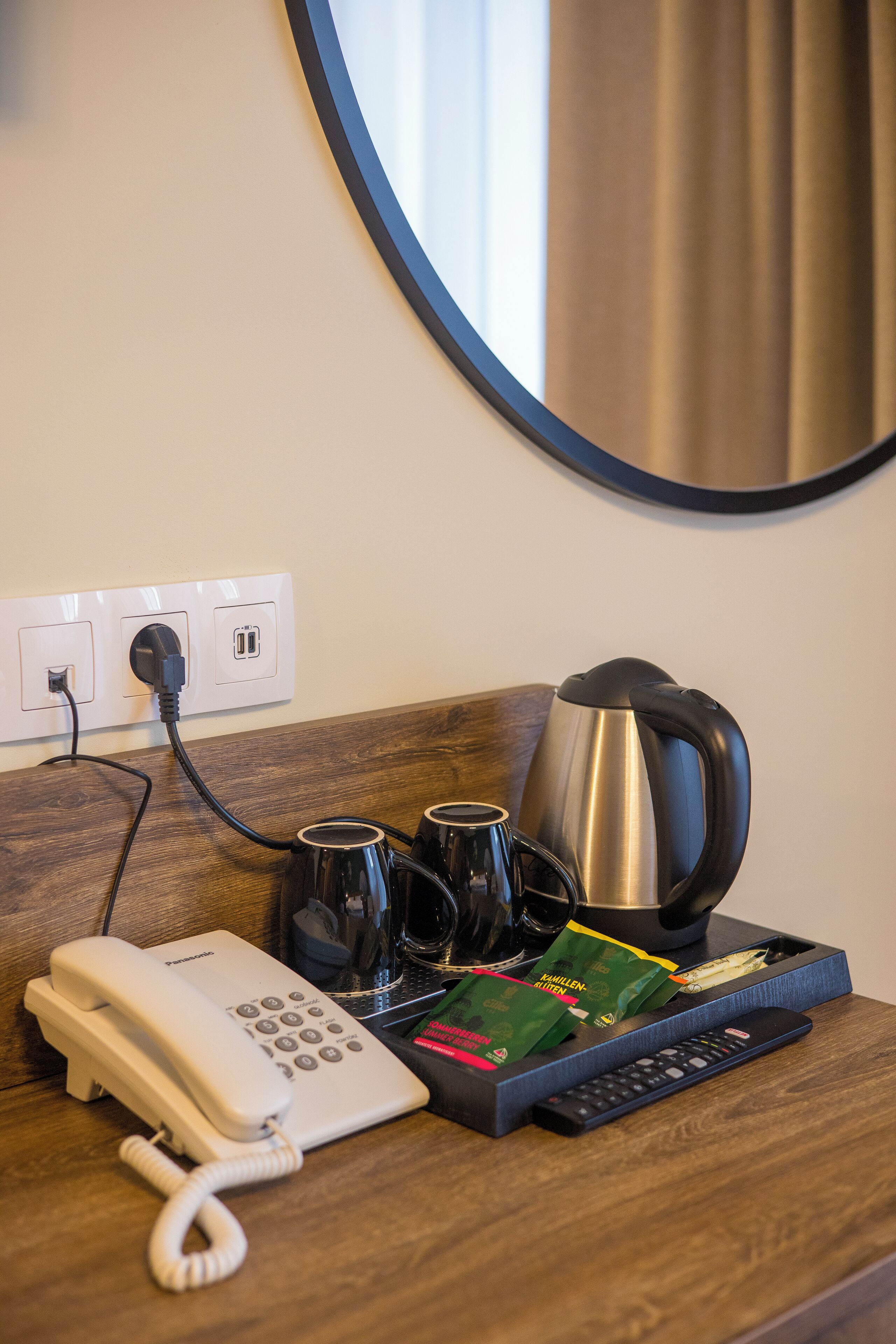 Hotelkamer met een houten bureau, een witte telefoon, een waterkoker, twee mokken, een dienblad met snacks en een afstandsbediening.