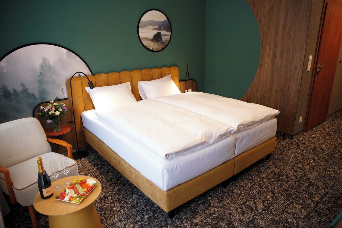 A neatly made bed with white linens, a wooden headboard, and a small table with a bottle and glasses beside it.