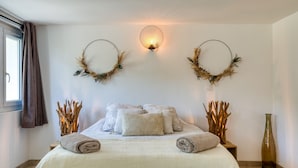Ein ordentlich bezogenes Bett mit weißer Decke und Kissen, eine an der Wand montierte Leuchte sowie zwei dekorative Kränze an beiden Seiten.