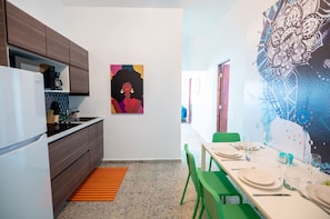 A compact kitchen with modern appliances, a dining area with green chairs, and a colorful abstract painting on the wall.