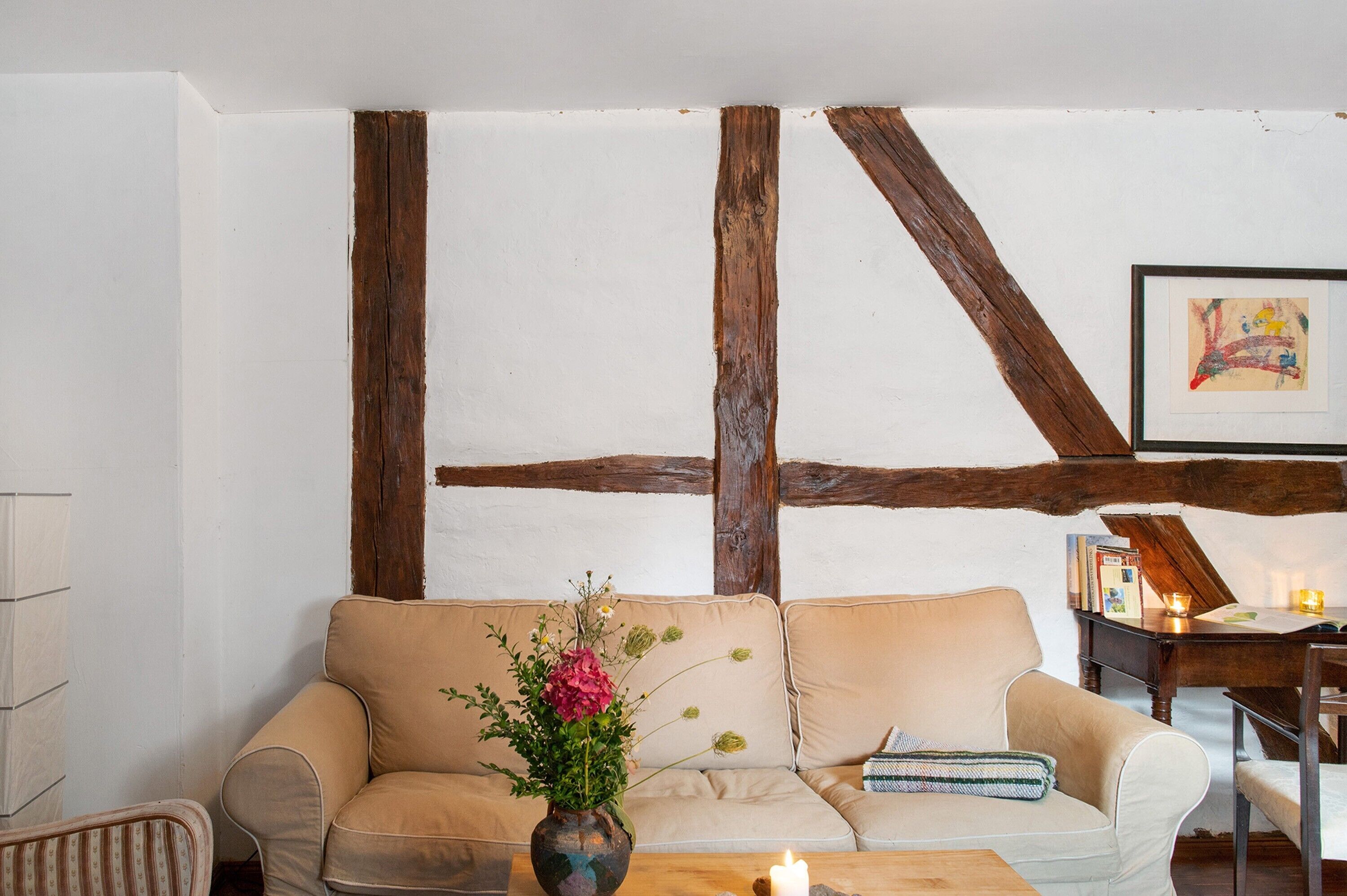 Ein Zimmer mit freiliegenden Holzbalken, einem beigen Sofa, einem kleinen Tisch mit Kerze und einem gerahmten Bild an der Wand.