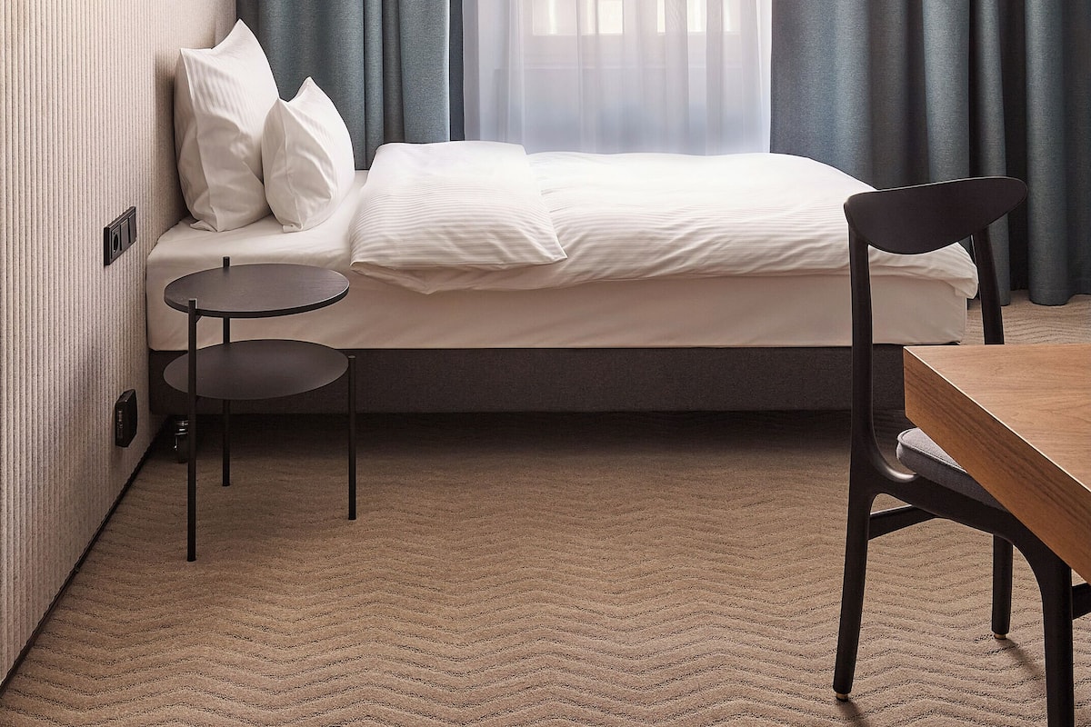A neatly arranged hotel room with a bed, two chairs, a small table, and a window with sheer curtains.