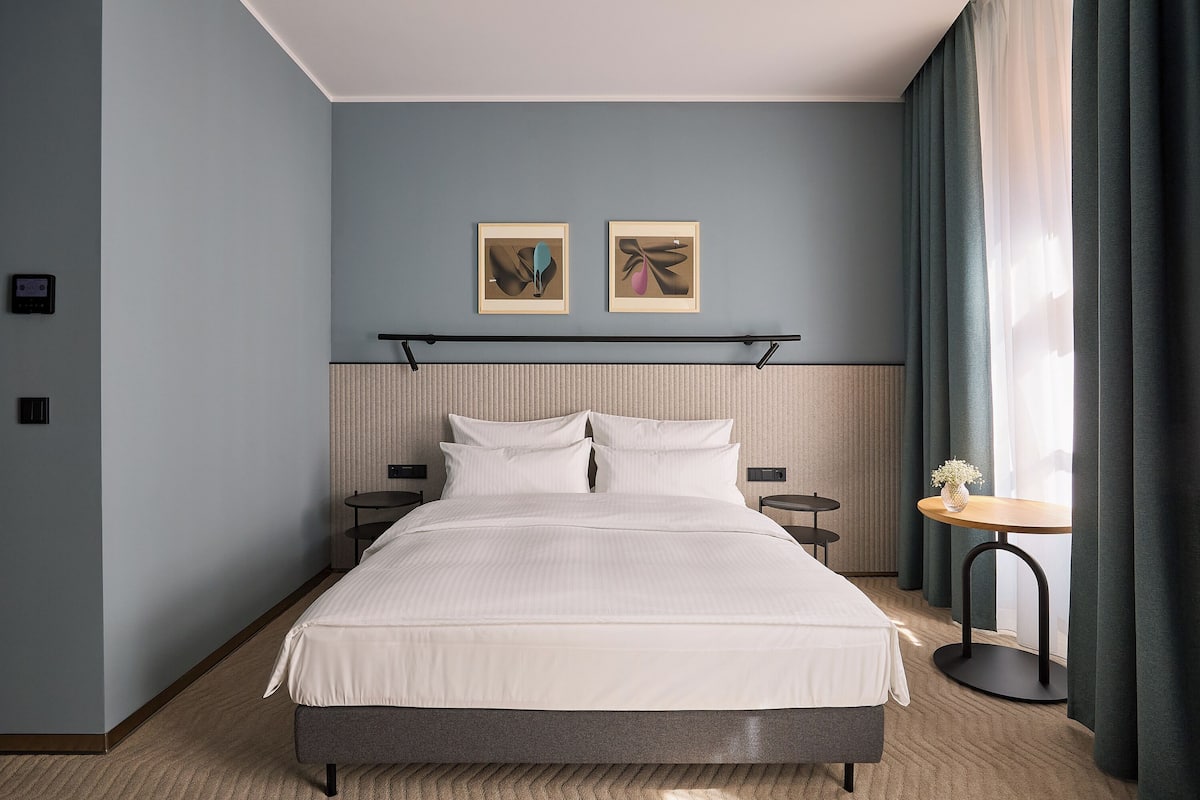 A hotel room with a large bed, two bedside tables, a round table with a vase, and two framed pictures on the wall.