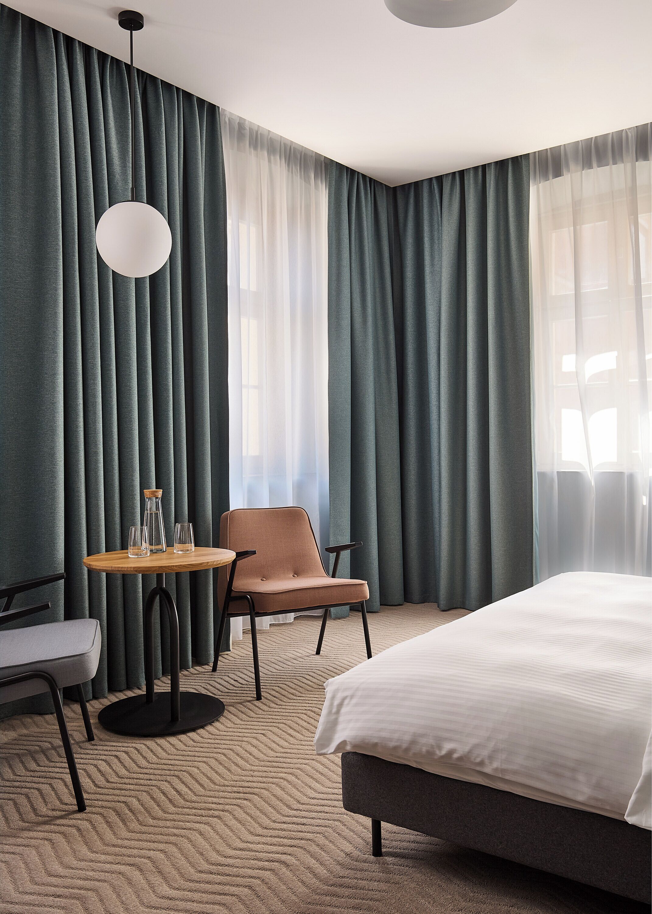 A hotel room with a bed, a chair, a table with glasses, and curtains.