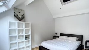 Une chambre avec un lit, une étagère blanche, une petite table et une œuvre d’art accrochée au mur.