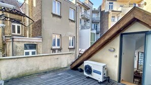 Une terrasse sur le toit avec une plateforme en bois, un climatiseur blanc et une porte vitrée donnant sur un espace intérieur.