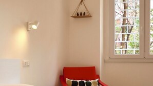 Une chambre d’hôtel avec un lit, un fauteuil rouge, une fenêtre donnant sur un paysage verdoyant et une étagère murale agrémentée d’objets décoratifs.