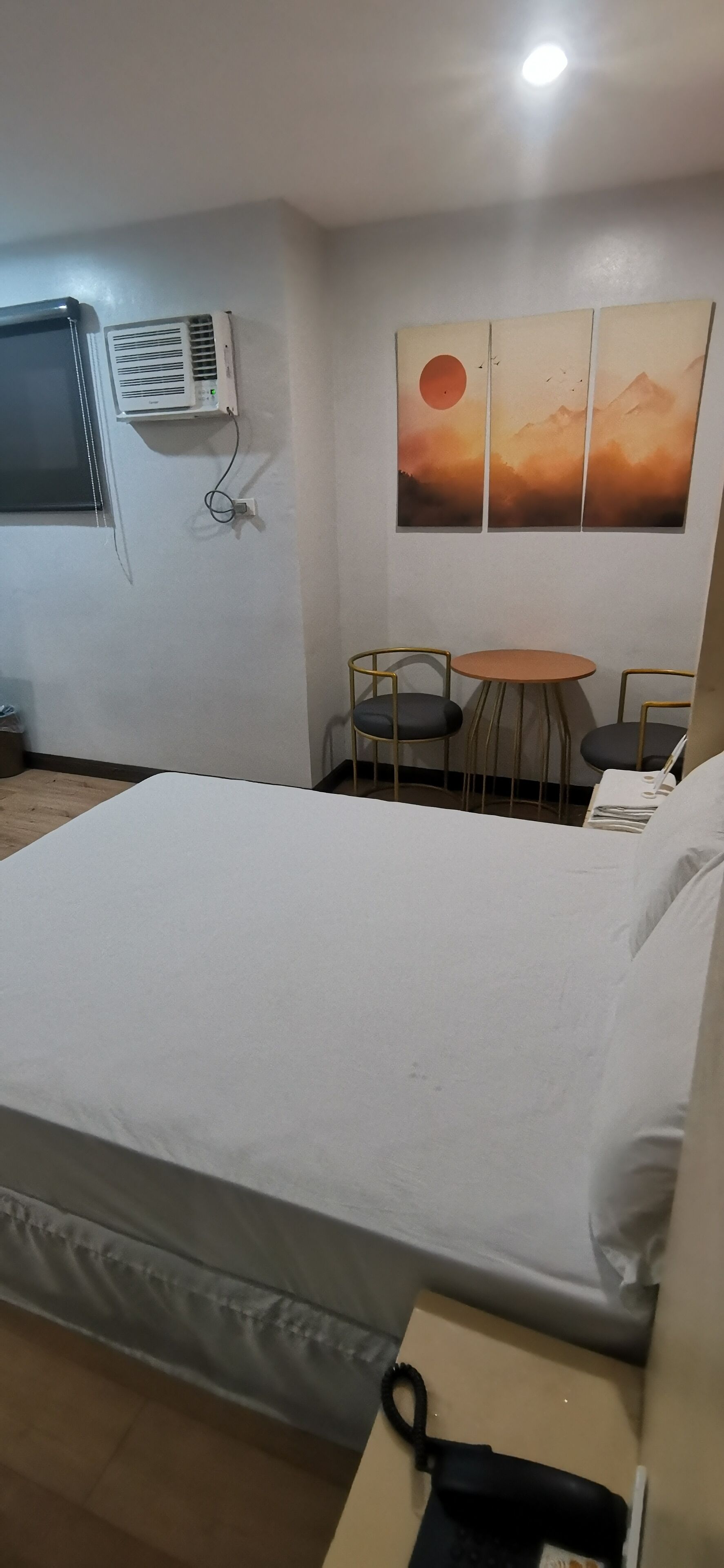 A hotel room with a bed, a small table with two chairs, an air conditioning unit, and a wall-mounted artwork depicting a sunset.