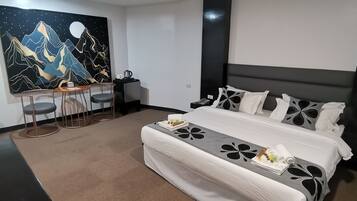 A modern hotel room with a large bed, a desk with chairs, a coffee maker, and a wall painting of mountains.