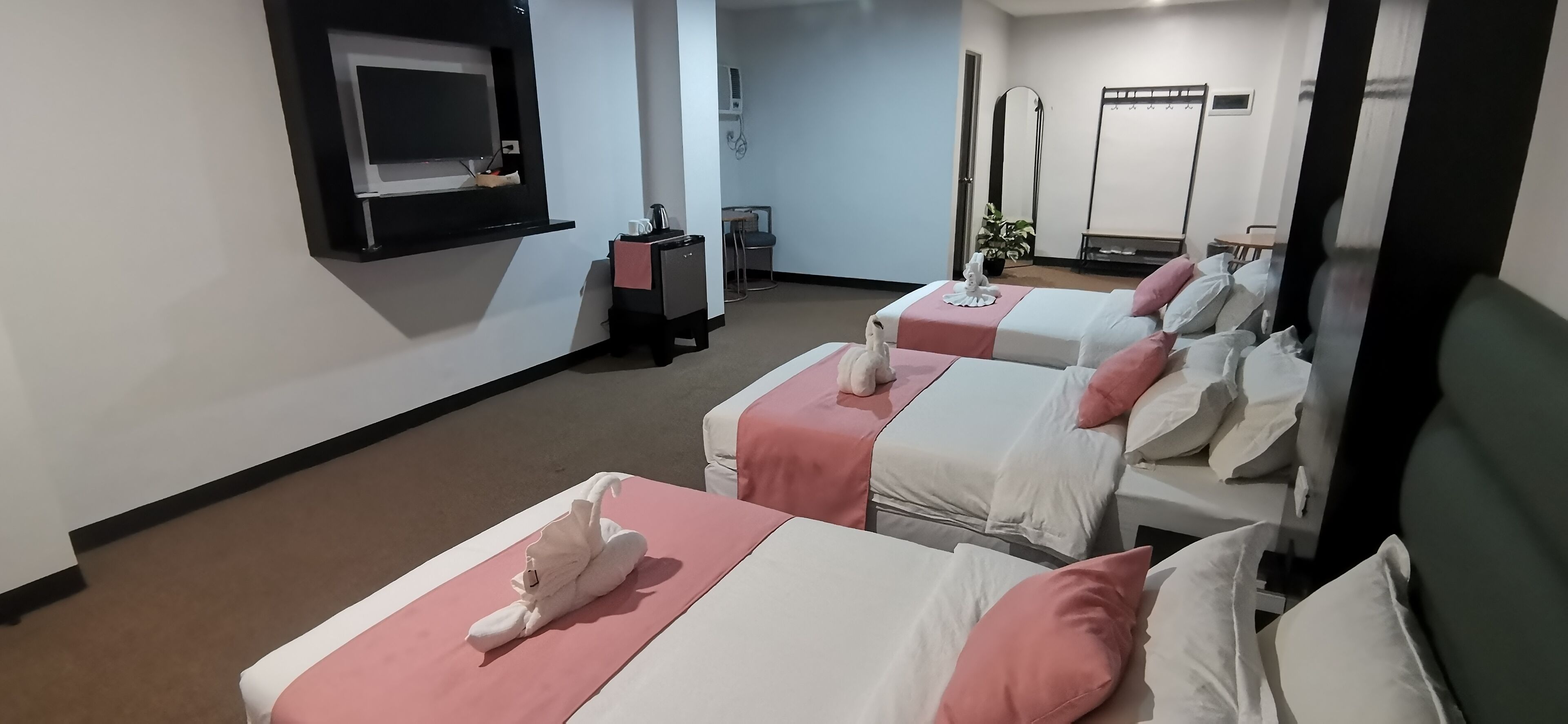 Habitación de hotel con tres camas, cada una con cubrecama rosa y blanco. Hay una pequeña mesa con un hervidor y una taza, y un televisor montado en la pared.