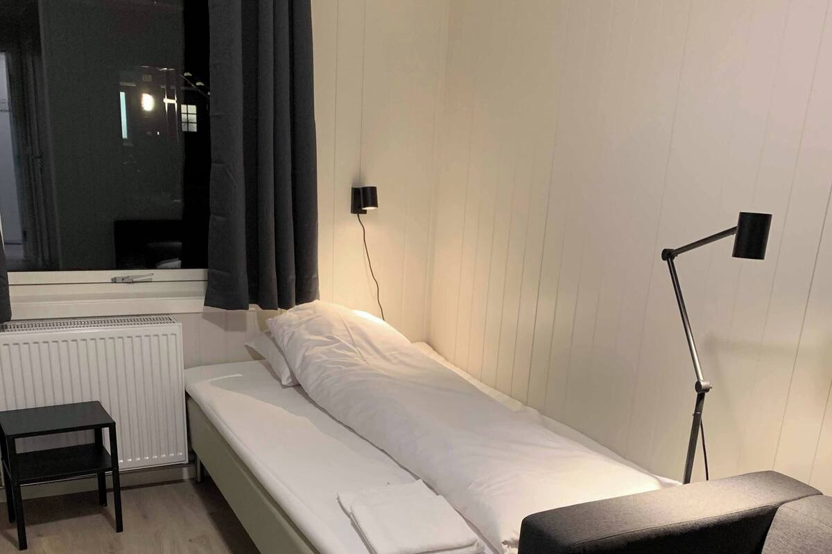 Un dormitorio ordenado con una cama individual, una mesita de noche, una silla, una lámpara de pie y un radiador.