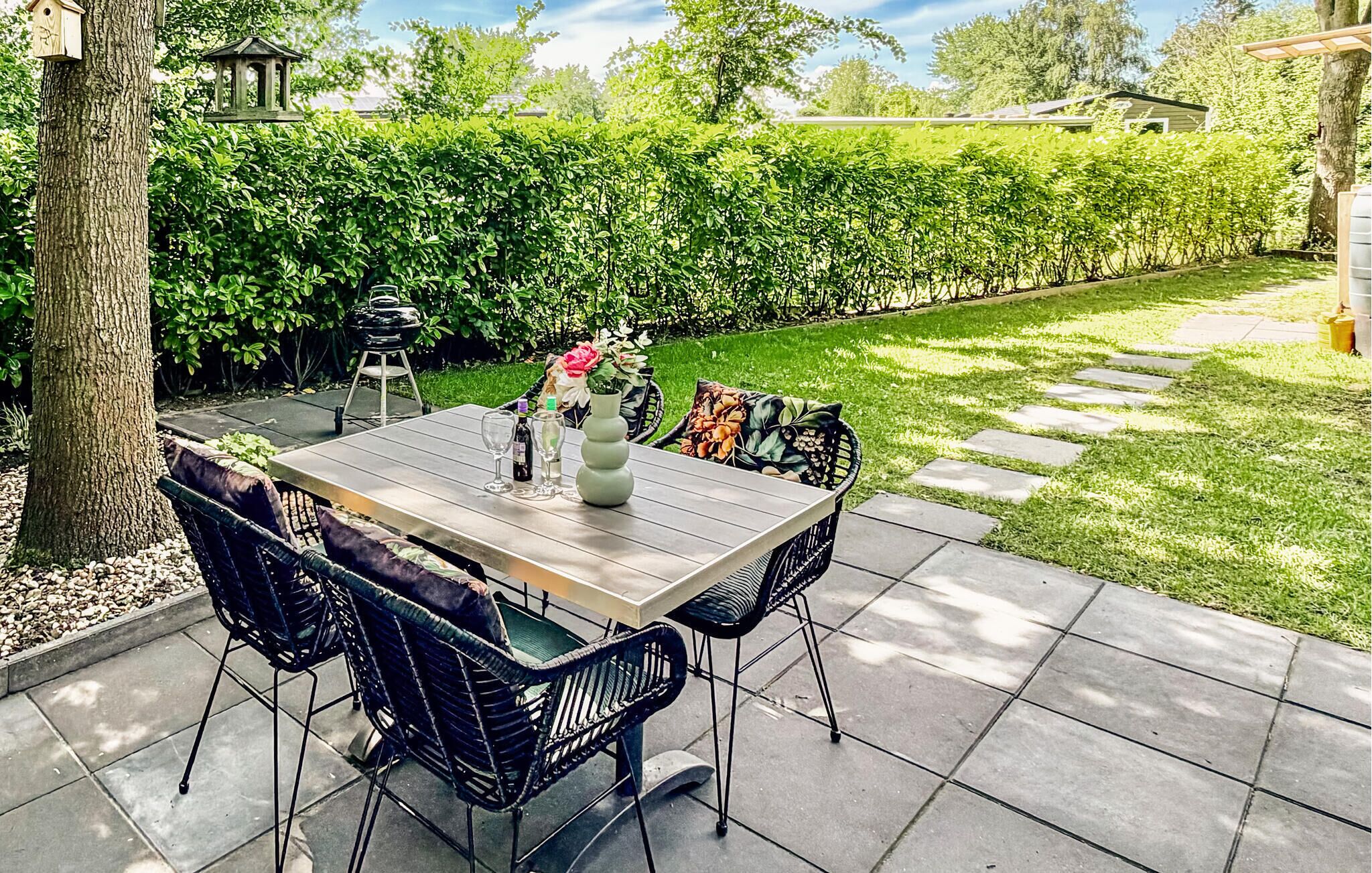 An outdoor patio with a wooden table and black wicker chairs, surrounded by a hedge and a paved walkway.