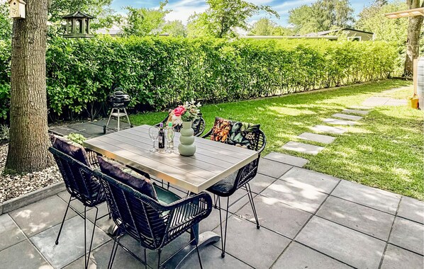 Un patio al aire libre con una mesa de madera y sillas de mimbre negro, rodeado de un seto y un camino pavimentado.