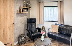 Una sala de estar con un sofá y un sillón de cuero negro, una mesa redonda de centro y una mesita auxiliar con un jarrón de flores. Se observa una ventana con cortinas, una puerta de madera y una estantería con objetos decorativos.
