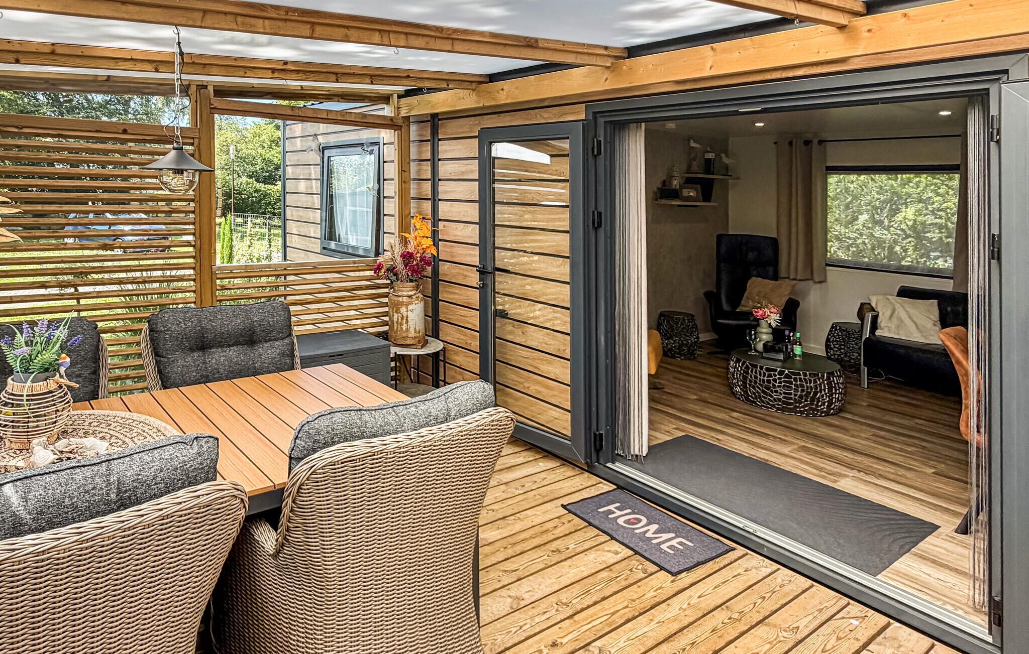 A cozy outdoor seating area with a wooden table and wicker chairs, a small side table with a vase, and a sliding glass door leading to an interior living space.