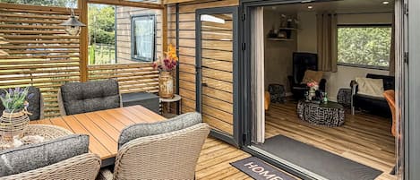 Een gezellig buiten zitgedeelte met een houten tafel en rieten stoelen, een bijzettafeltje met een vaas, en een glazen schuindeur die leidt naar een leefruimte binnenshuis.