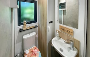 Une salle de bain avec des toilettes, un lavabo et une fenêtre. Un panneau est accroché au mur avec l’inscription suivante : « Vivez par la foi, grandissez en grâce, agissez avec amour. »