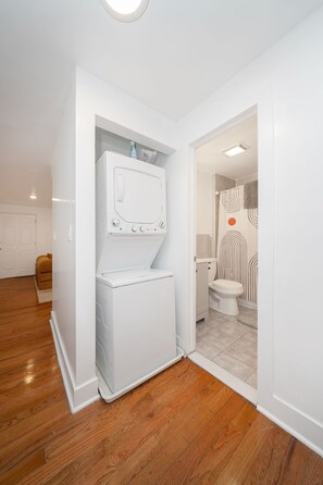 Un cuarto de lavado con lavadora y secadora, piso de madera y una puerta blanca que da a un baño con cortina de ducha a rayas.