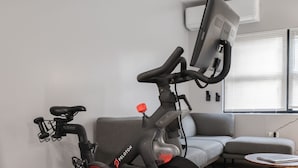 Una bicicleta de ejercicio Peloton en una sala de estar con un sofá gris, una mesa de centro redonda y una ventana con persianas.