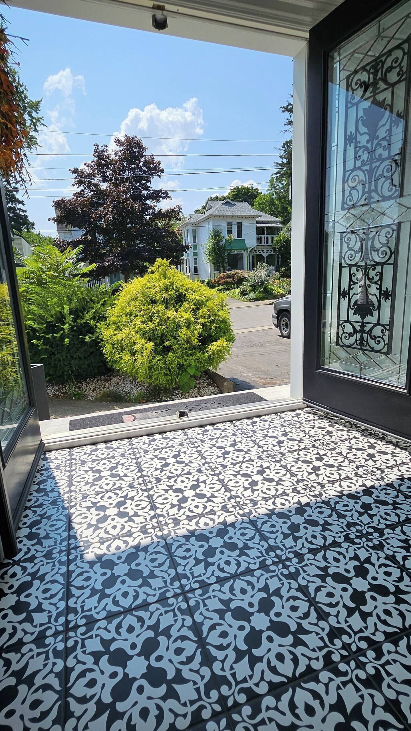 Une vue par une fenêtre sur un sol carrelé à motifs, un buisson vert et une maison à l’extérieur blanche et aux bordures vertes.