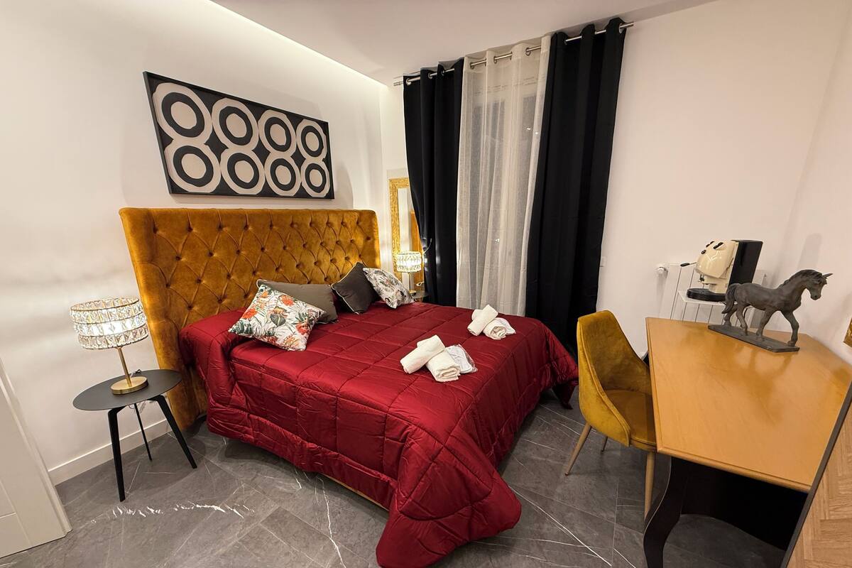 A hotel room with a tufted headboard, a red bedspread, a small round table with a lamp, a chair, and a desk with a horse figurine.