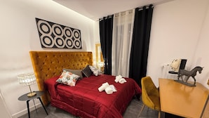 Una habitación de hotel con cabecera capitonada, colcha roja, una pequeña mesa redonda con una lámpara, una silla y un escritorio con una figurina de caballo.