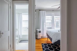 A bedroom with a bed, a window with curtains, a radiator, and a view of the city.