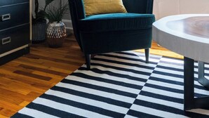 A modern living room with a teal armchair, a round wooden table, a black and white striped rug, a floor lamp, and a large window with curtains.