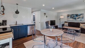 A modern kitchen with a dishwasher, a dining table with chairs, a sofa, a TV, and a painting on the wall.