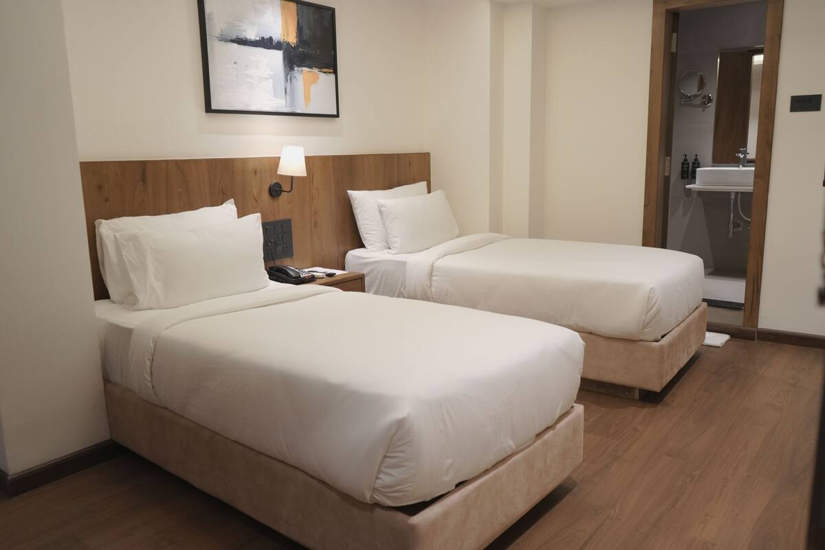 Ein Hotelzimmer mit zwei Einzelbetten, einem Holzkopfteil, einem Nachttisch mit Lampe und einem durch eine offene Tür einsehbaren Badezimmer.