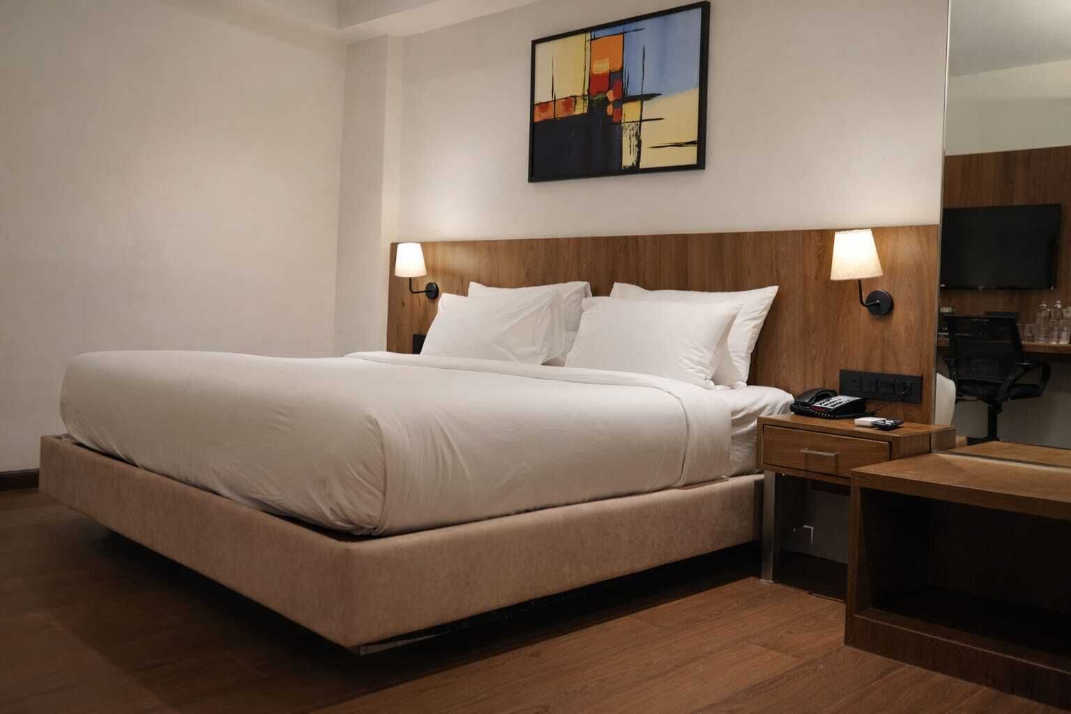 A hotel room with a large bed, two bedside tables, a desk, a chair, a TV, and a painting on the wall.