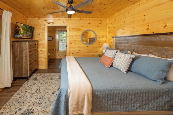 Un dormitorio de cabaña de madera con cama, ventilador de techo, espejo y televisor sobre un tocador.