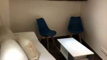 Une petite pièce chaleureuse, avec un lit, deux chaises bleues, une table blanche et des poutres en bois au plafond.
