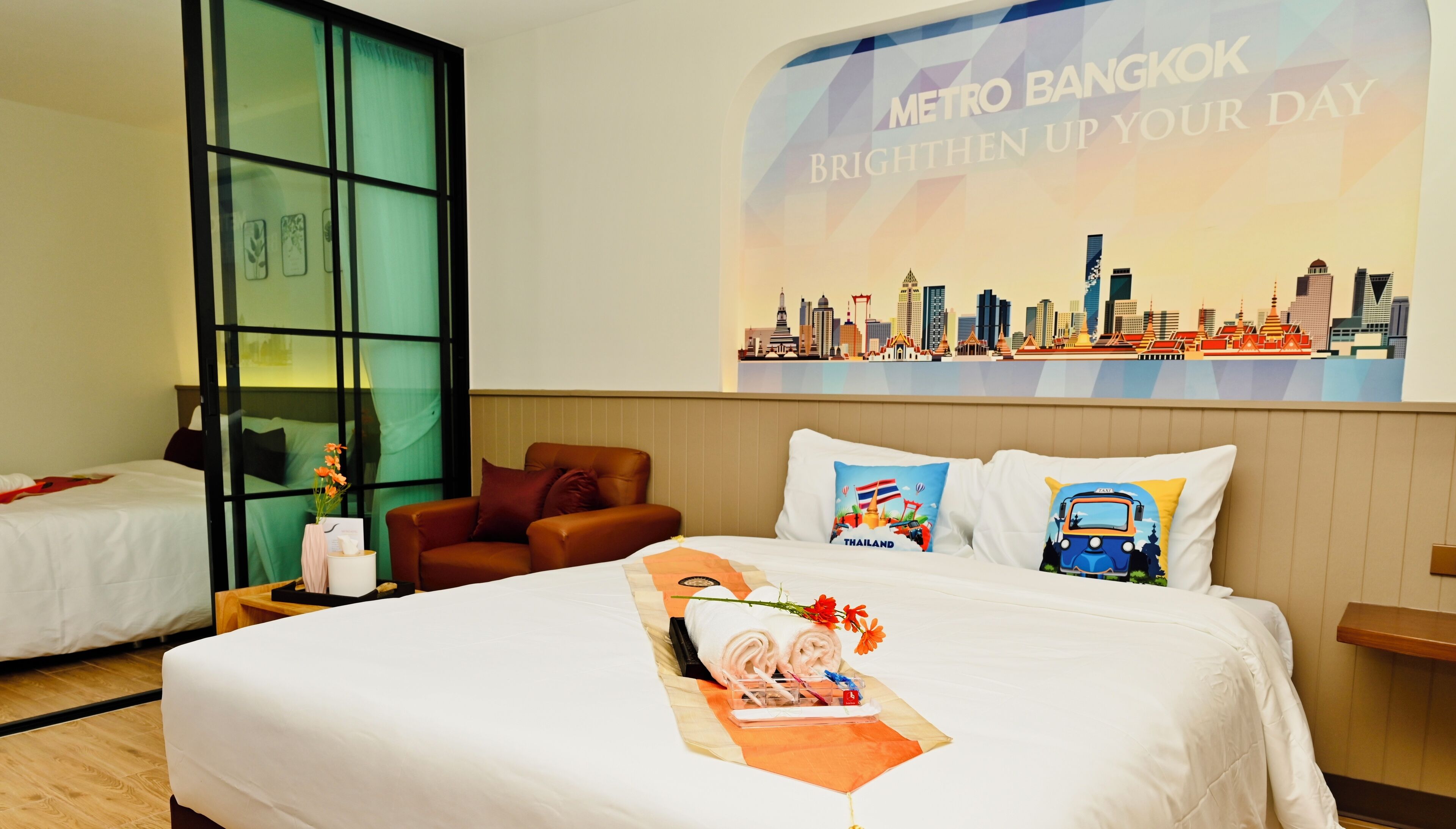 Une chambre d’hôtel avec un lit, deux oreillers ornés de motifs sur le thème de la Thaïlande, un fauteuil en cuir marron, une table de chevet et un mur décoré d’une affiche représentant un paysage urbain.