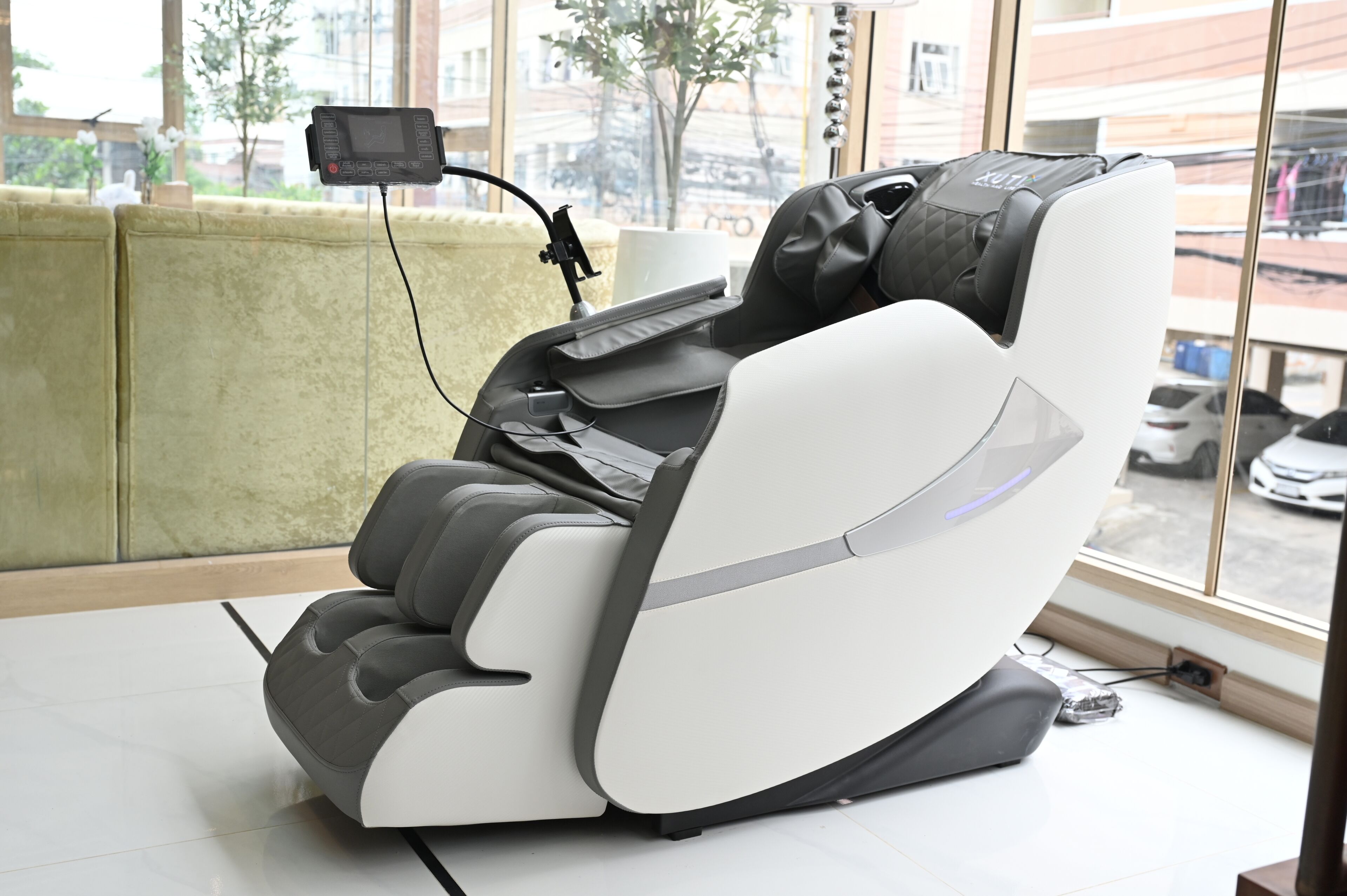 A massage chair with a control panel and a charging cable, positioned near a window with a view of the street.