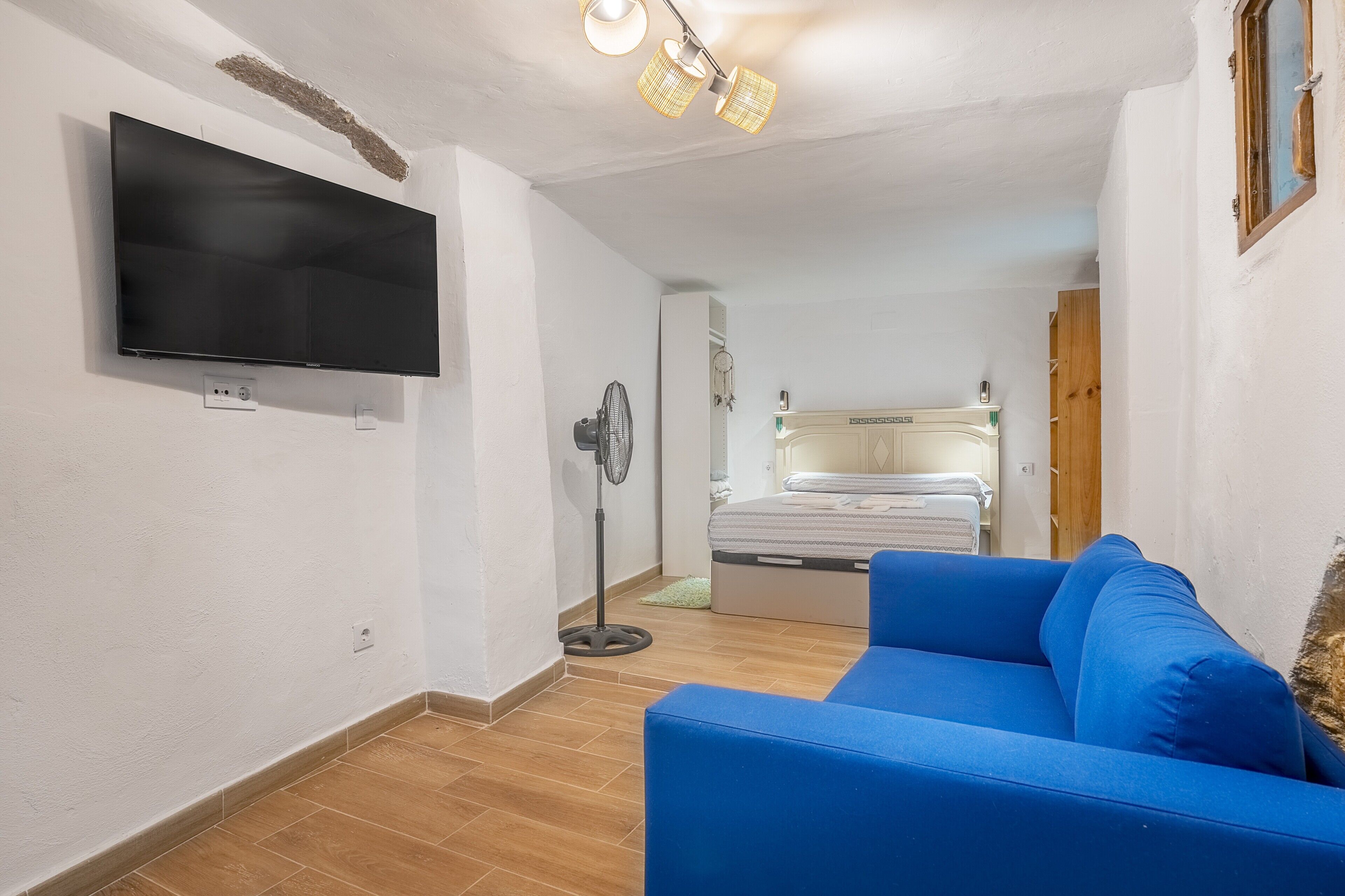 A room with a flat-screen TV mounted on the wall, a blue sofa, a bed with a white headboard, and a ceiling fan.