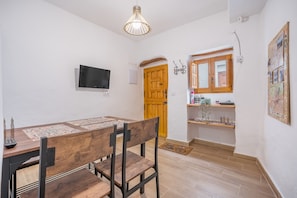 A dining area with a wooden table and chairs, a television mounted on the wall, a wooden door, and a shelf with various items.