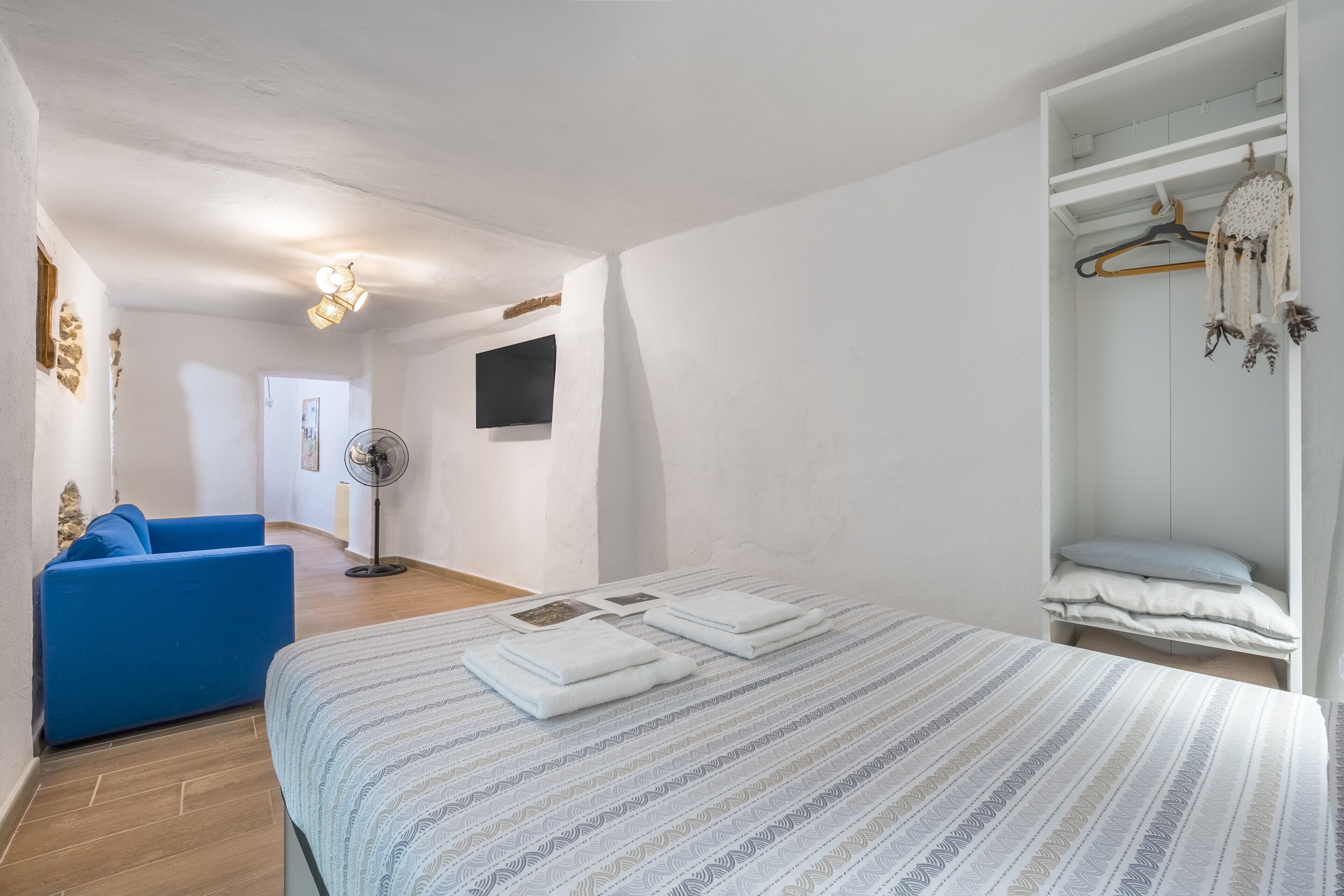 Un dormitorio con cama, una silla azul, un armario y un ventilador.