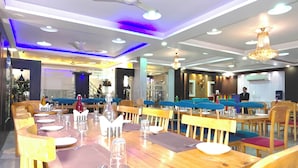 A restaurant with wooden tables and blue cushioned chairs, a ceiling fan, and a chandelier. - Hotel Aman Suites at IGI by Arch Group (New Delhi)