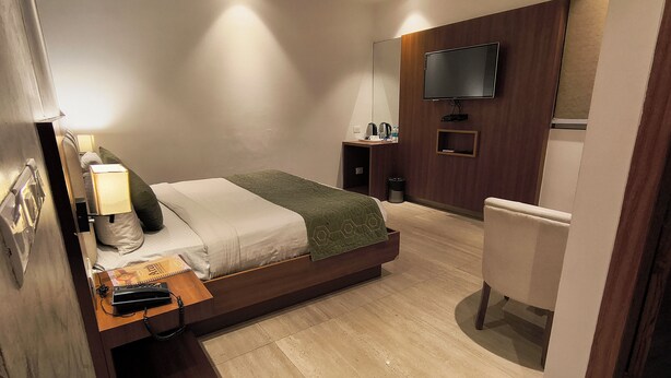 A hotel room with a bed, bedside table, lamp, TV, and a small desk with a chair. - Hotel Aman Suites at IGI by Arch Group (New Delhi)