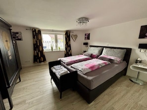A bedroom with a bed, bedside tables, a dresser, a window with curtains, and a chandelier.