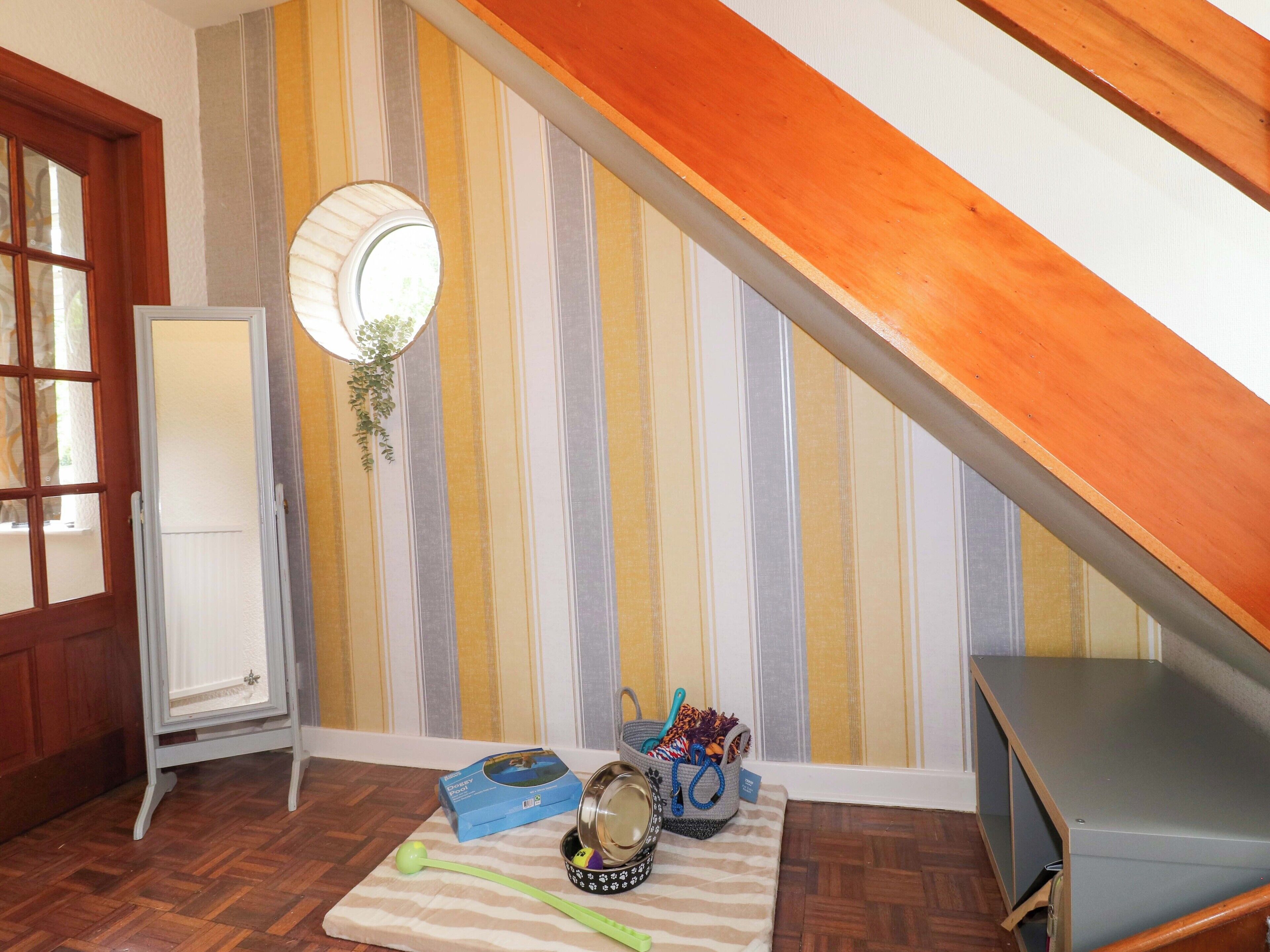 A room with striped wallpaper, a wooden sloped ceiling, a mirror, a pet bowl, a leash, and a basket.