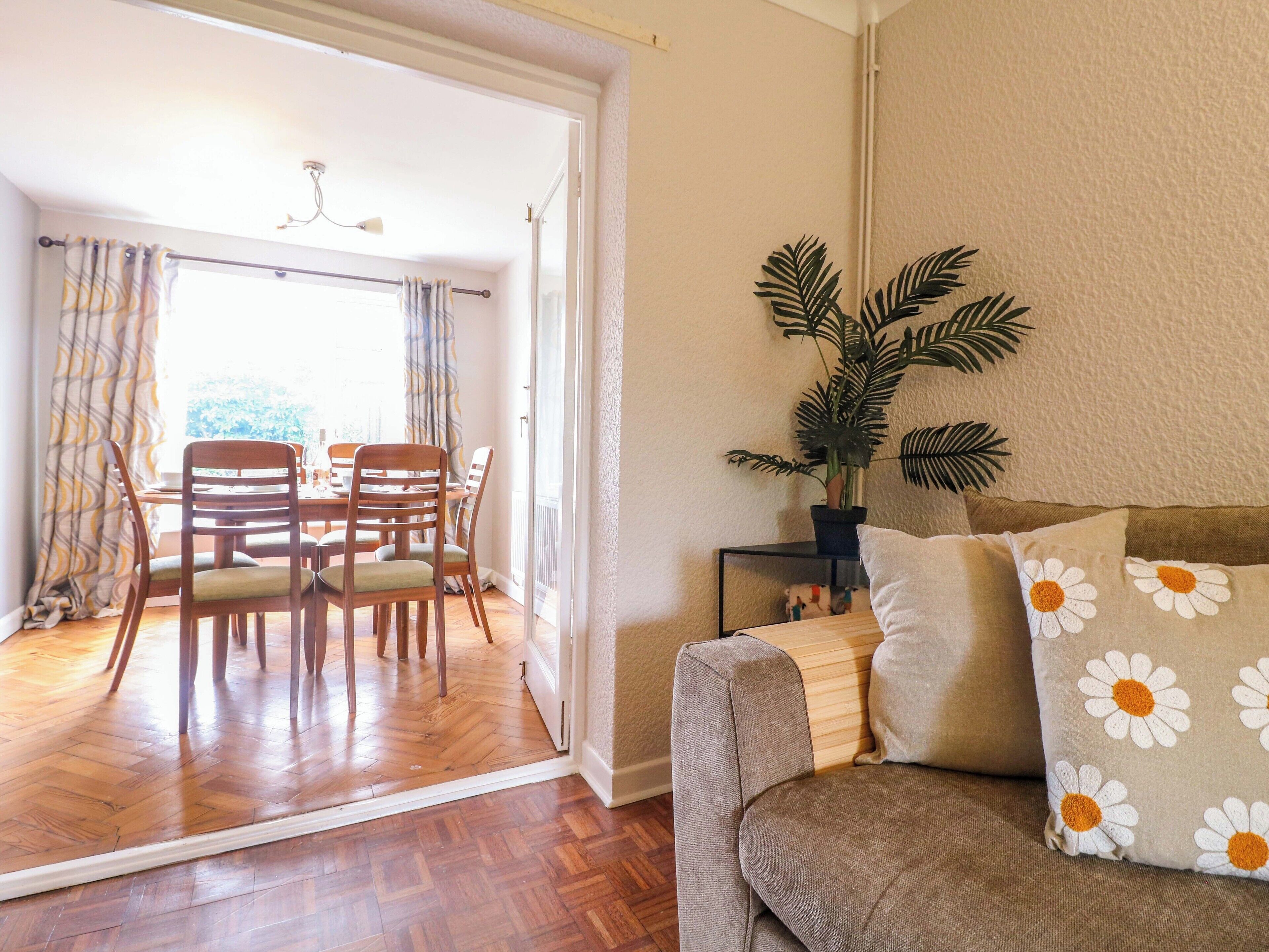 A living room with a wooden floor, a sofa with daisy-patterned cushions, a dining table with chairs, and a potted plant.