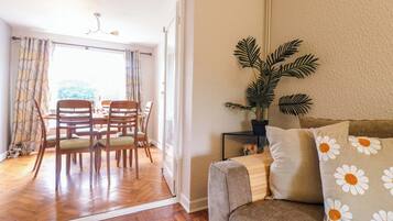 A living room with a wooden floor, a sofa with daisy-patterned cushions, a dining table with chairs, and a potted plant.
