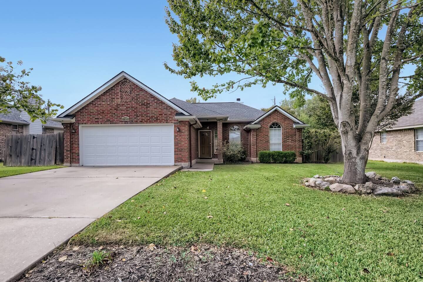 A two-story brick house with a white garage door, a front yard with a tree, and a wooden fence.