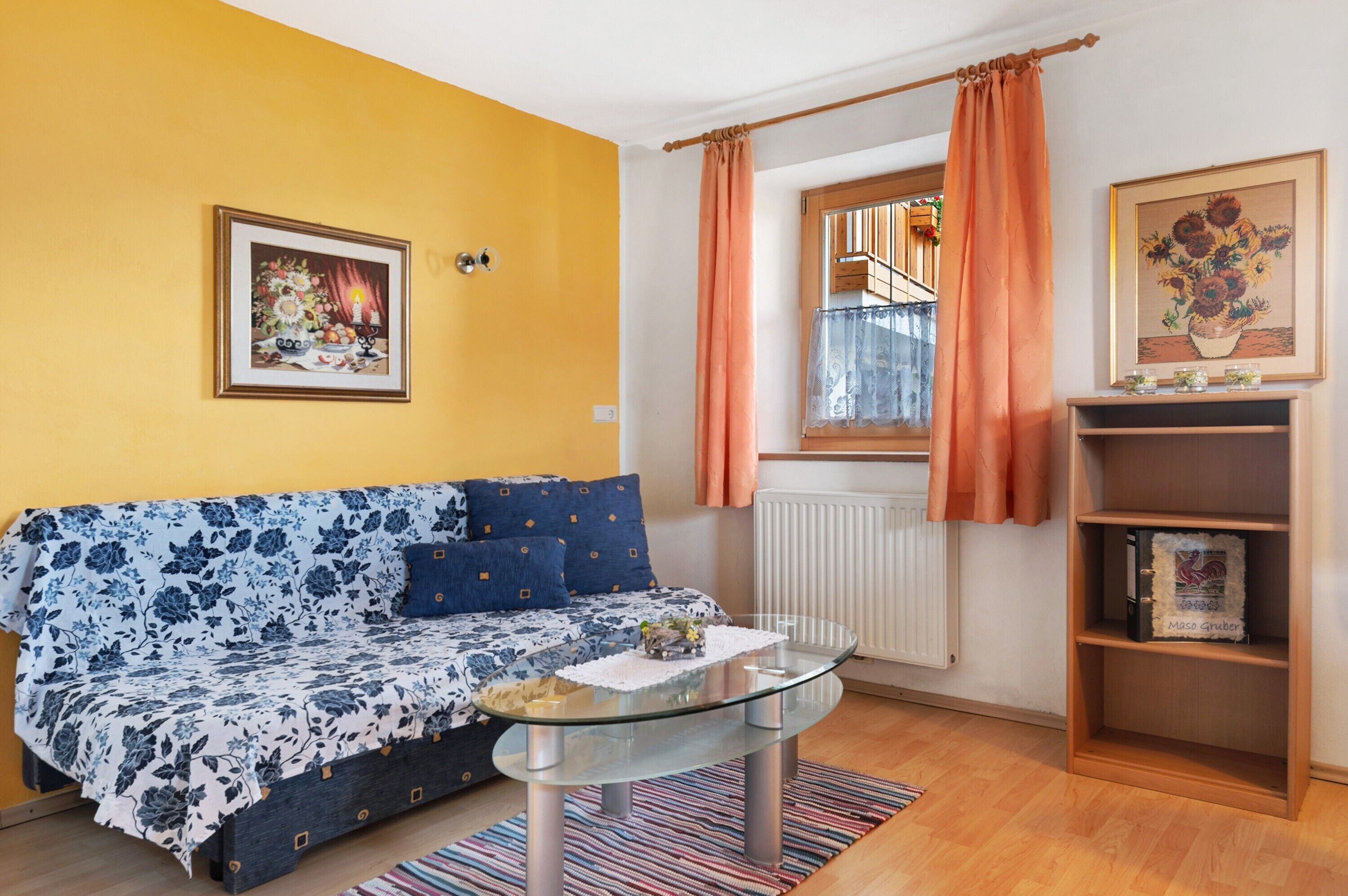 Una sala de estar con un sofá, una mesa de centro de vidrio, una estantería, una ventana con cortinas y un radiador.