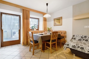 A dining area with a table, chairs, a window with curtains, a hanging light fixture, and a bed with a floral bedspread.