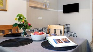Uma área de refeições com uma mesa posta para dois, uma tigela de frutas, um vaso com flores e um livro.