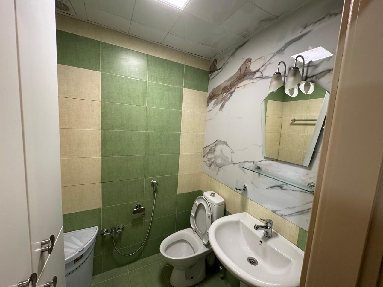 A bathroom with a toilet, bidet, and sink, green and beige tiles, and a marble backsplash.